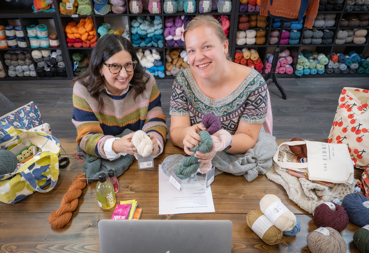 Two women sitting at a table with yarn and knitting materials in a store setting.