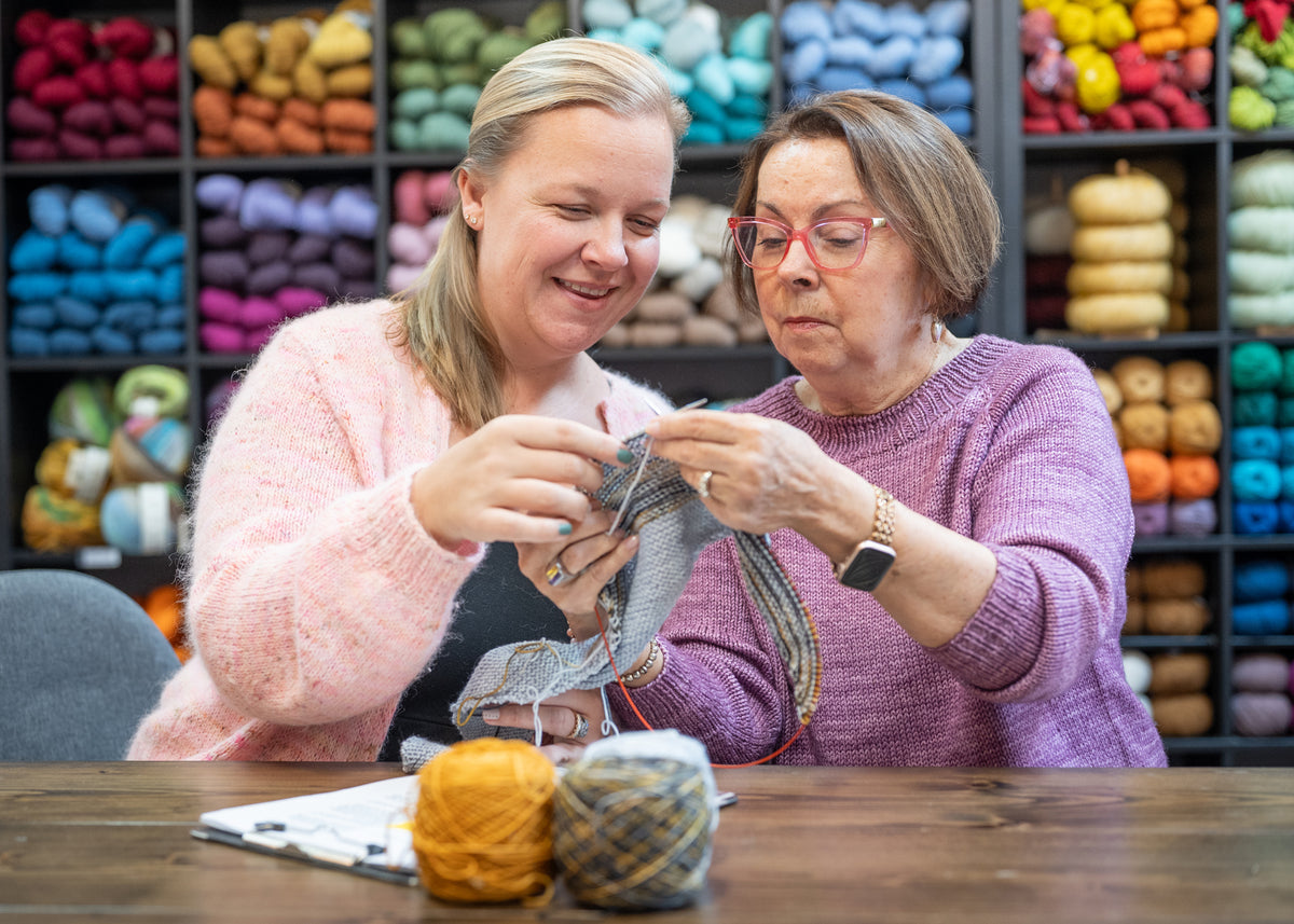 Two women knitting together in a yarn store with colorful yarn on shelves in the background.