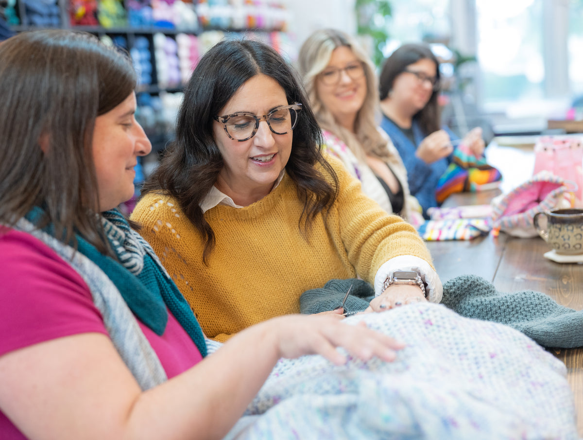 Two women sitting at a table in a craft store, looking at yarn and knitting materials.