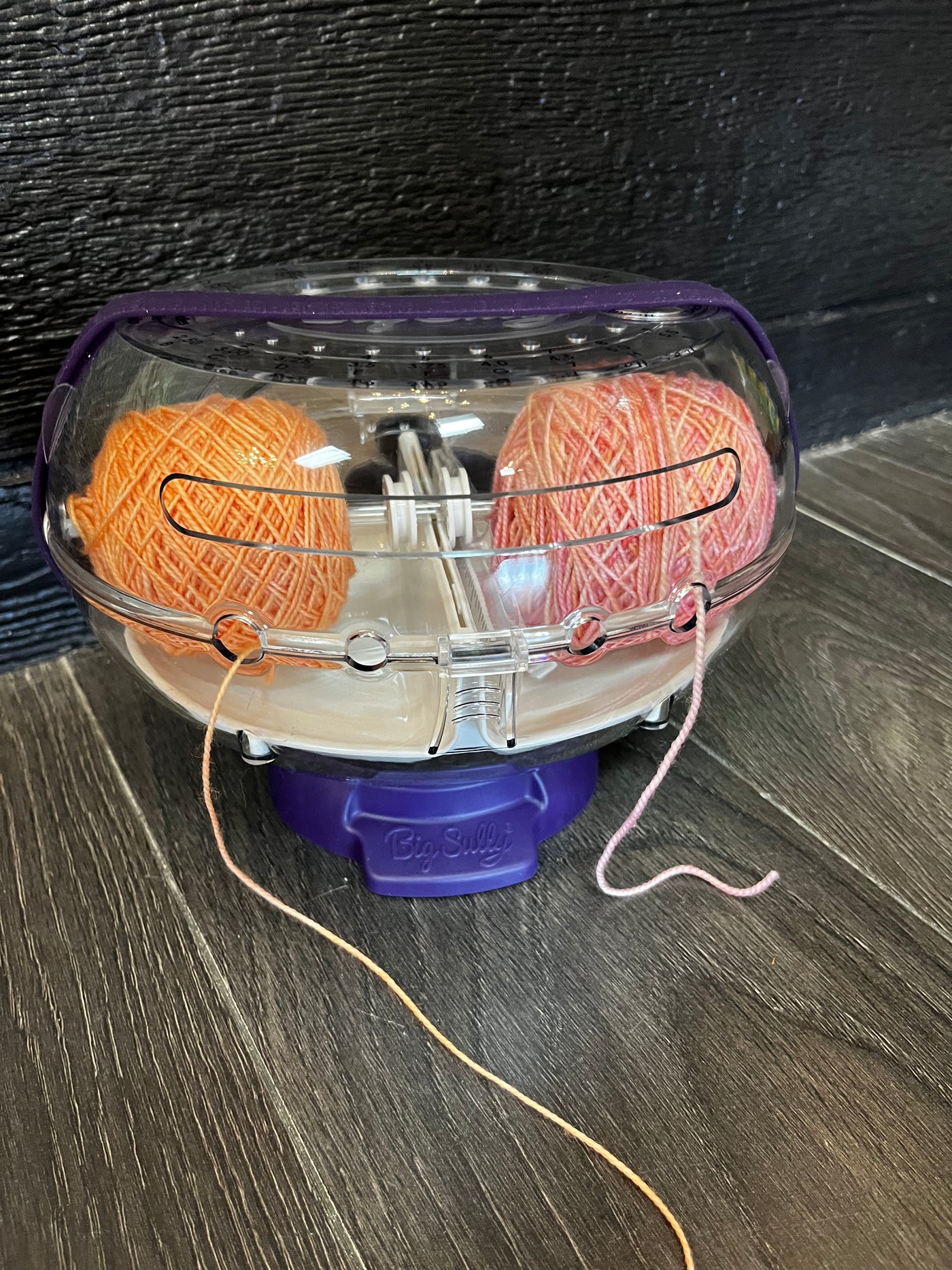A double yarn ball holder made of clear plastic with a purple base that reads "Big Sully" on a wood floor. It is holding peach cakes of yarn. 