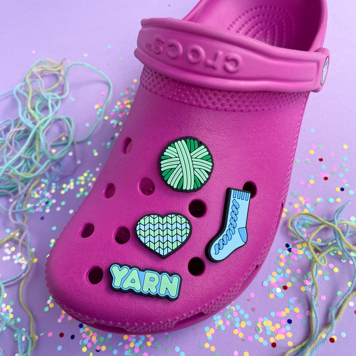 A hot pink Croc shoe with charms on it shaped like a yarn ball, heart, sock, and the word yarn. The charms are shades of mint green and blue. 