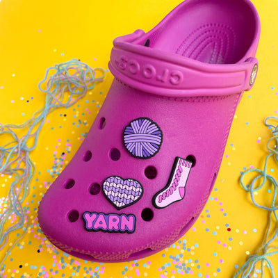 A hot pink shoe with PVC charms shaped like a heart, yarn ball, sock, and the word yarn in shades of pink and purple.
