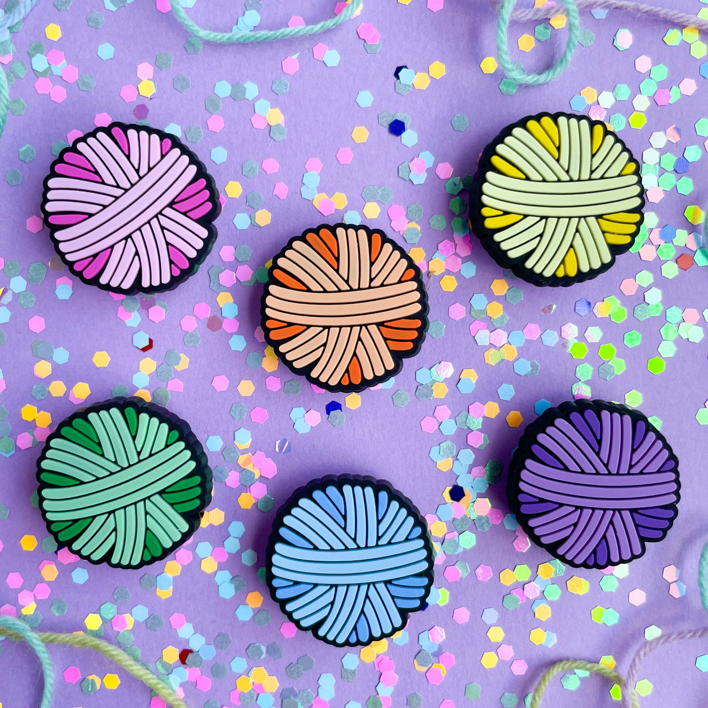 PVC charms shaped like yarn balls in pink, orange, yellow, mint, blue, and purple on a lavender background covered in confetti. 
