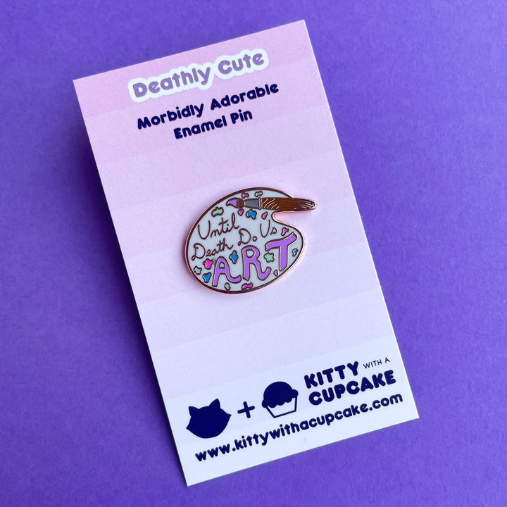 An enamel pin shaped like an artist's palette with a paint brush that reads "until death do us art" on a pink card.