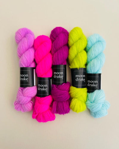 A group of bright neon pink skeins of yarn with lime and sky blue skeins. The yarn is very fluffy with a visible fuzzy texture due to the brushed cashmere fiber content.