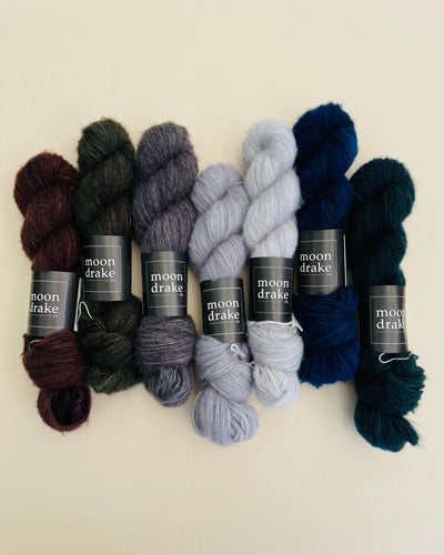 Skeins of cashmere yarn lined up together in shades of cool grey, deep blue, dark grey, and burgundy. The yarn is very fluffy and fuzzy.