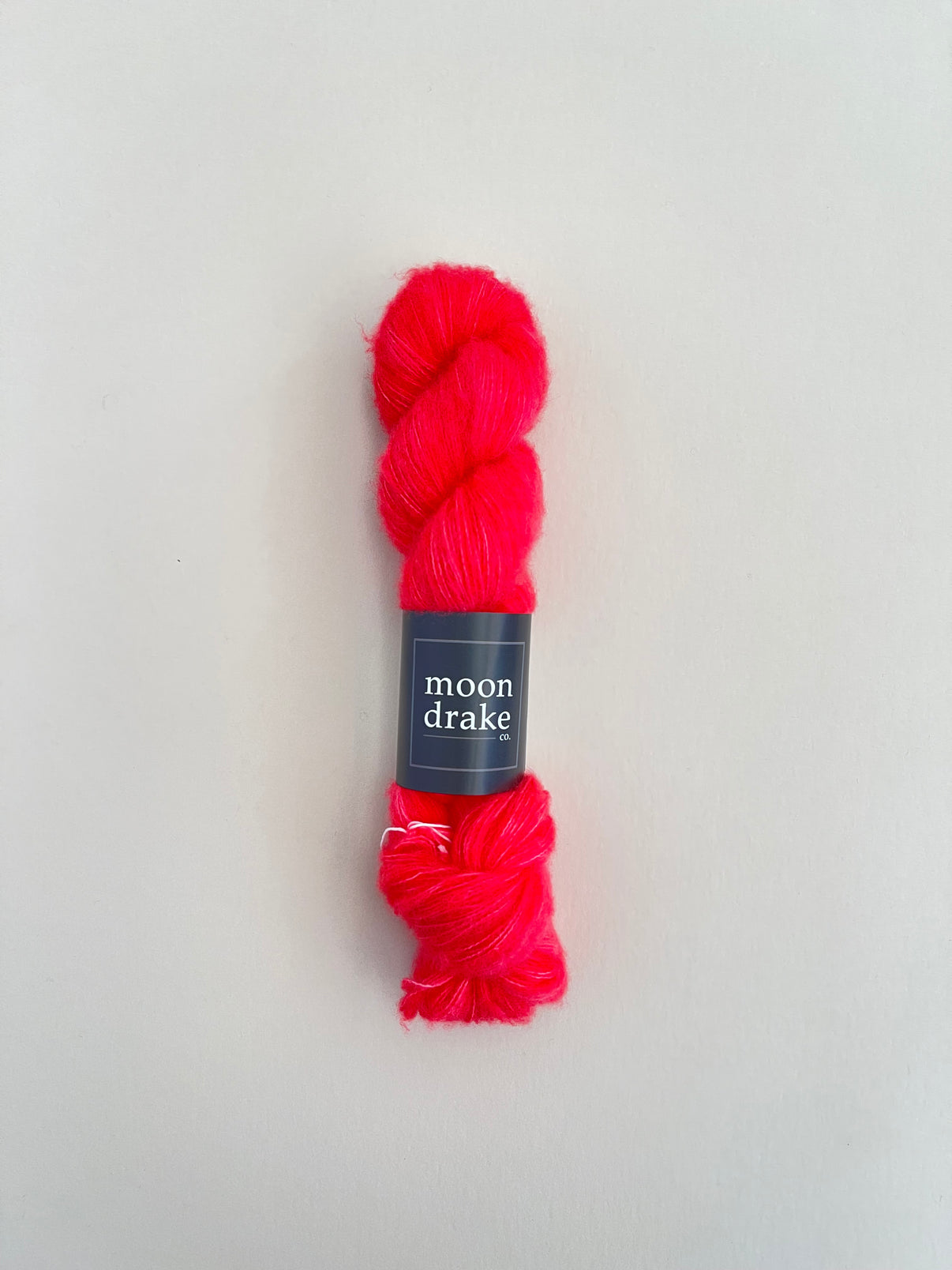 A very bright coral colored skein of yarn with a black label that reads "Moondrake Co". The yarn is very fluffy and floofy.