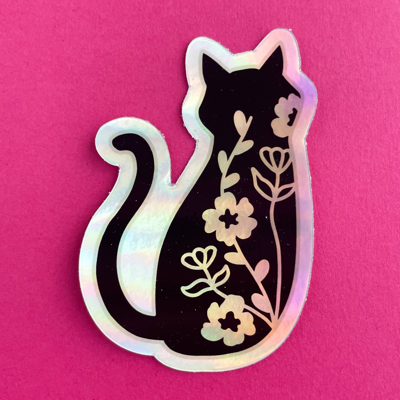 A holographic sticker in the shape of a silhouette of a black cat. There are floral vines along the back of the cat. The sticker is on a hot pink background.